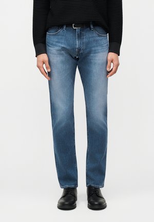 Mężczyzna ubrany w niebieskie proste dżinsy, czarny pasek, czarny prążkowany sweter i czarne sznurowane buty, stojący na tle jednolitego jasnego tła.