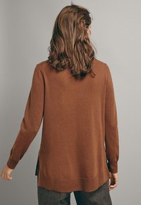 Personne avec des cheveux bruns ondulés portant un long pull marron et un pantalon à carreaux foncé, vue de dos sur un fond uni.