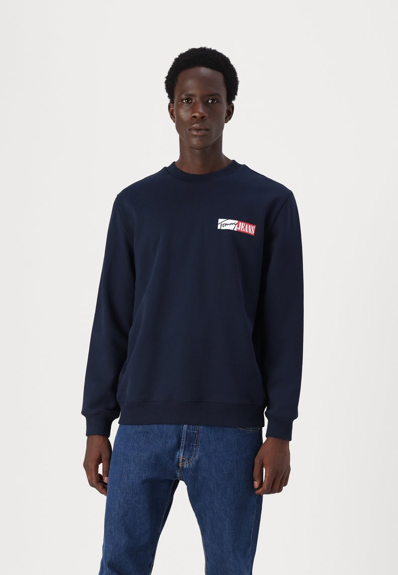 Marineblå crewneck sweatshirt med ribbede mansjetter, med en logo i rødt og hvitt på venstre bryst. Mykt, teksturert stoff.