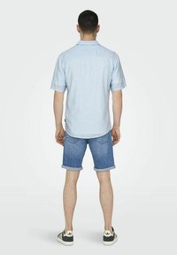 Hellblaues kurzärmliges Hemd mit vertikalen Streifen, kombiniert mit hochgekrempelten Jeansshorts. Das Modell trägt schwarz-weiße Sneaker.