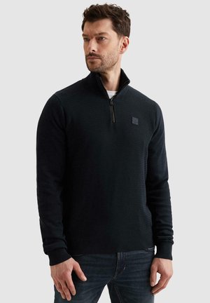 Man met kort haar en baard, gekleed in een zwarte, getextureerde trui met rits en donkere spijkerbroek, kijkt opzij tegen een effen achtergrond.