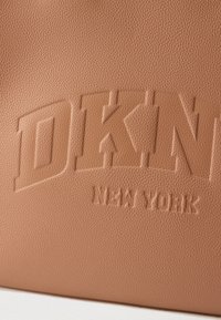 Logo embossé « DKNY New York » sur une surface en cuir texturé couleur noisette, mettant en valeur les détails de la marque et la qualité du matériau.