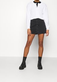 Cardigan bianco peloso con bottoni, abbinato a una gonna di jeans nera con bottoni frontali, e stivaletti neri con lacci.