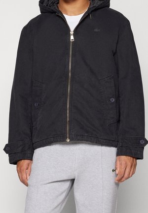 Mann trägt eine schwarze Zip-up-Kapuzenjacke mit knopflappentaschen und graue Jogginghosen mit kleinem grünem Logo am Oberschenkel.