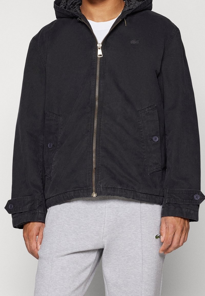 Homme portant une veste noire à capuche zippée avec des poches à rabat boutonnées et un pantalon de survêtement gris avec un petit logo vert sur la cuisse.
