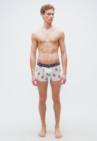 Νεαρός άνδρας που στέκεται ξυπόλυτος φορώντας λευκά μπόξερ με σχέδιο αρκουδάκι και μπλε ζώνη μέσης Ralph Lauren.