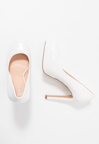 Bílé lakované kožené lodičky s kulatou špičkou, elegantním designem a zúženým stiletto podpatkem, který má kontrastní hnědou akcentaci na základně.