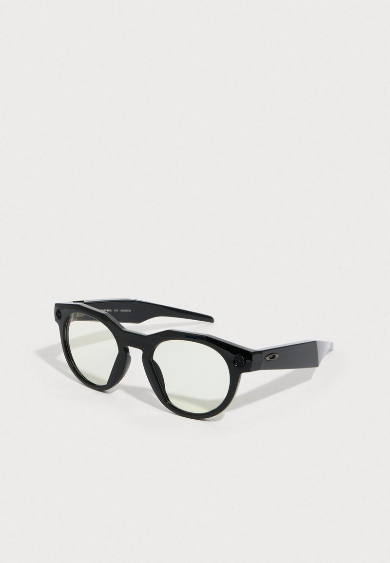 Lunettes de soleil noires avec une forme arrondie, des verres teintés verts, des montures noires angulaires et des accents latéraux. Surface texturée avec un détail en métal.