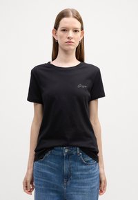 Juodas medvilninis T-shirt su trumpomis rankovėmis ir apvalia kaklo išpjova. Apvalkala puošia subtilus baltas siuvinėtas logotipas ant krūtinės. Derinami su mėlynomis džinsinėmis kelnėmis.