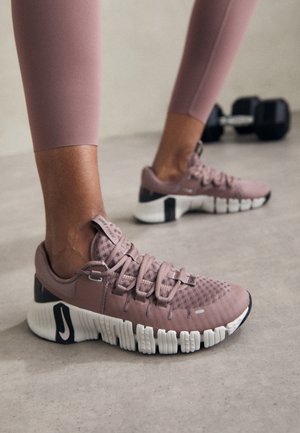 Personne portant des chaussures de sport mauves et des leggings assortis, debout sur un sol clair avec un haltère en arrière-plan.
