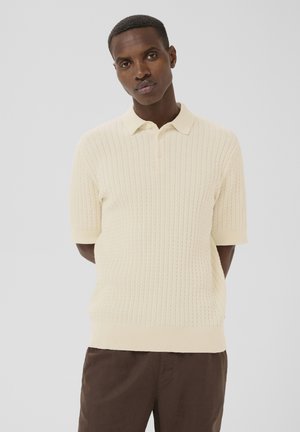 Uomo con capelli corti che indossa una polo a maniche corte color crema in maglia strutturata e pantaloni marroni, in piedi con le mani dietro la schiena su sfondo grigio.