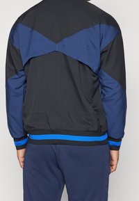Black and navy athletic jacket featuring a contrasting blue stripe at the hem, with a unique chevron design on the back.