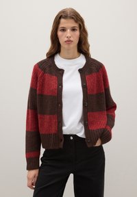 Cardigan à rayures rouges et brunes avec une texture côtelée, un col rond et des fermetures à boutons. Porté par-dessus une chemise blanche et un pantalon noir.