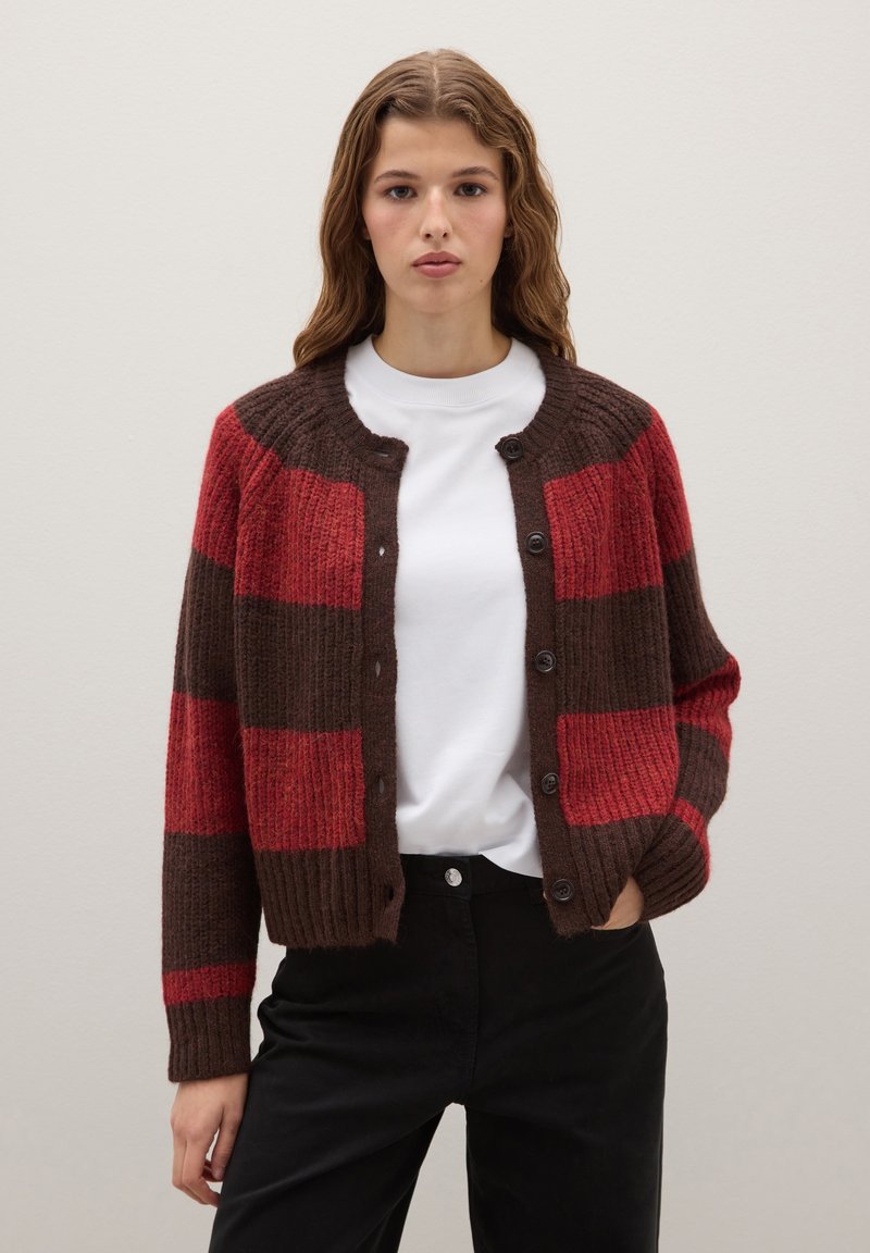 Cardigan à rayures rouges et brunes avec une texture côtelée, un col rond et des fermetures à boutons. Porté par-dessus une chemise blanche et un pantalon noir.