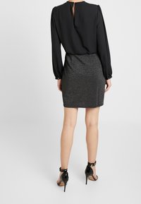 Černá blůza s dlouhým rukávem a nabíranými rameni, spárovaná s úzkou, strukturovanou černou mini sukni. Lesklé černé podpatky dokončují outfit.