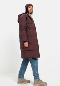 Gevulde kastanjebruine jas met een capuchon, ritssluiting en zijzakken. Gepaard met lichtblauwe jeans en beige veterschoenen. Zachte textuur.