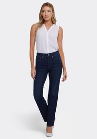 Witte mouwloze blouse met V-hals, gecombineerd met donkerblauwe high-waisted jeans; model staat met handen langs haar zij. Witte hakken.