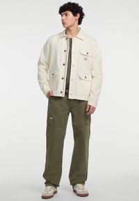 Weiße Denimjacke mit zwei Brusttaschen und silbernen Druckknöpfen, kombiniert mit olivgrünen Cargo-Hosen und beigen Sneakers mit braunen Akzenten.