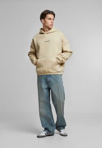 Beige hoodie met een voorzak, met een klein zwart logo. Draagt het met loszittende, vervaagde blauwe jeans en grijze sneakers.