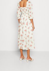 Vestido maxi floral en crema claro con rosas pastel; mangas abullonadas, espalda fruncida, parte inferior en capas. Combinado con sandalias de tacón de color claro.