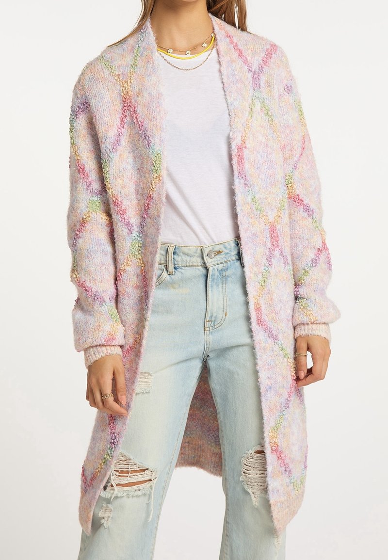 Femme portant un long cardigan pastel à motif losanges, sur un t-shirt blanc et un jean bleu clair déchiré.
