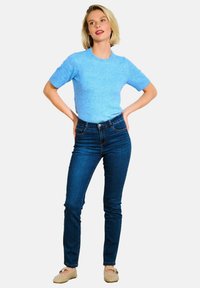 Světle modrý top s krátkým rukávem zkombinovaný s tmavě modrými skinny džíny a béžovými nazouvacími botami s výřezy. Modelka pózuje s rukama na bocích.