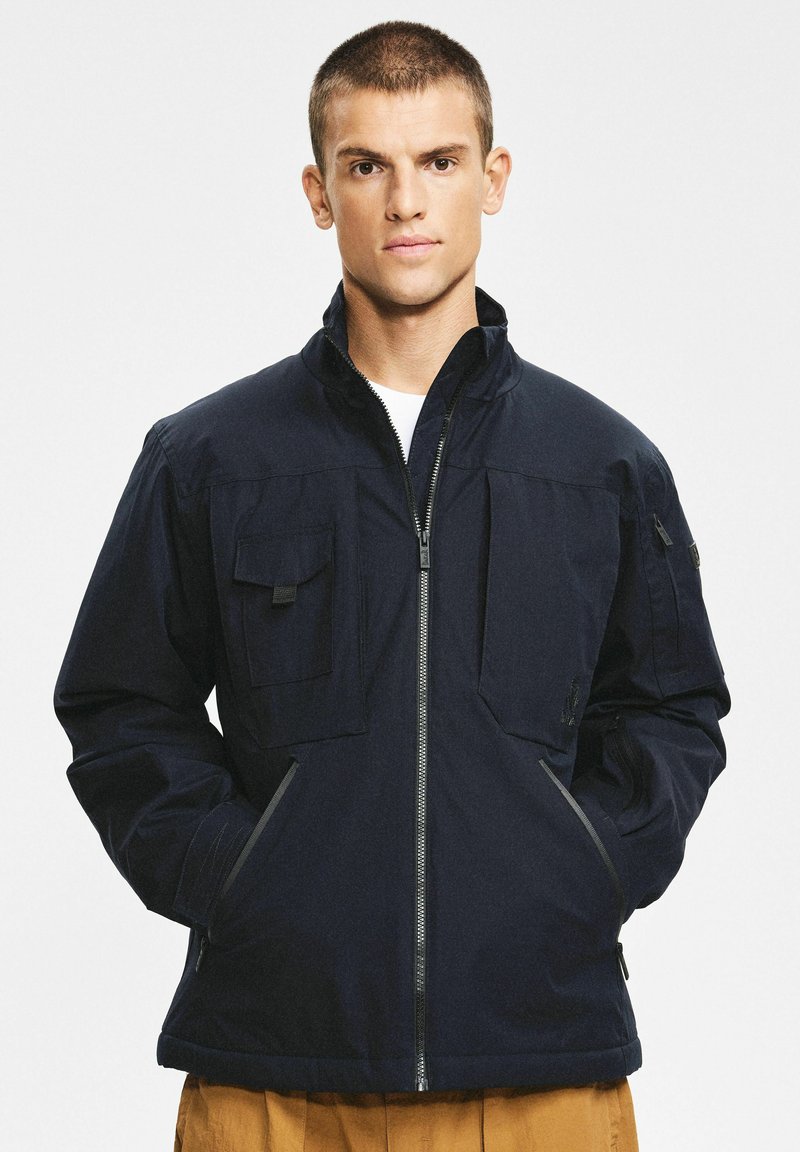 Granatowa kurtka na zamek z dwoma kieszeniami na piersi, kieszeniami bocznymi oraz prążkowanym kołnierzem. Charakteryzuje się gładką teksturą i casualowym stylem.