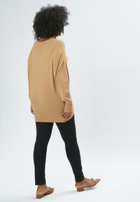 Beige oversized striktrøje med ribbede manchetter, parret med sorte tætsiddende bukser og brune slip-on sko med dekorativt accent.