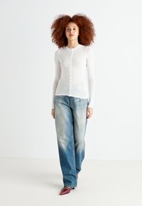 Weißes Langarmoberteil mit Knopfleiste, kombiniert mit hellblauen weiten Jeans. Die Model trägt bordeauxfarbene spitze Schuhe. Schlichter Hintergrund.