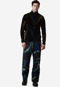 Giacca in pile nera con un motivo sottile; abbinata a pantaloni a motivi camouflage in blu scuro, verde e accenti gialli. Stivali robusti completano l'outfit.