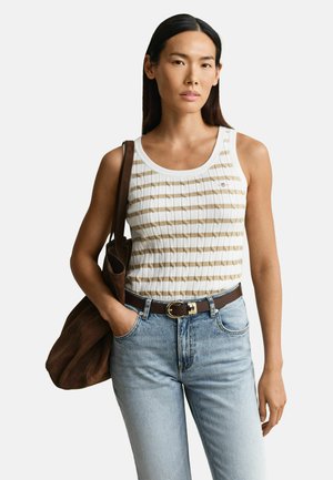 Vrouw met lang donker haar, gekleed in een beige-wit gestreepte mouwloze top, lichtblauwe spijkerbroek, bruine riem, en met een bruine suède tas over haar schouder.