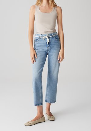 Frau trägt ein beiges Tanktop, hellblaue Jeans mit hoher Taille und weißem Seilgürtel sowie beige flache Schuhe, steht vor einem schlichten Hintergrund.