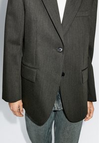 Blazer su misura grigio scuro con rever a punta, due tasche frontali e chiusura con un solo bottone, abbinato a jeans azzurri.