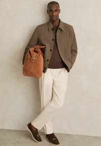 Brun yderjakke, mørk sweater, cremefarvede bukser; holder en tan ruskinds taske med spænde. Brune ruskinds sko med lys sål, står mod en neutral baggrund.
