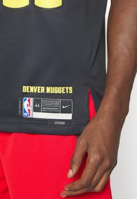Czarna koszulka koszykarska z żółtym napisem "DENVER NUGGETS". Posiada logo NBA i metkę Nike, boczne rozcięcie oraz przewiewną siateczkę. Widać czerwone spodenki.