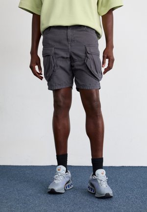 Szare szorty cargo z wieloma bocznymi kieszeniami, w połączeniu z jasnoszarymi trampkami i czarnymi skarpetkami. Tło jest gładkie, białe.