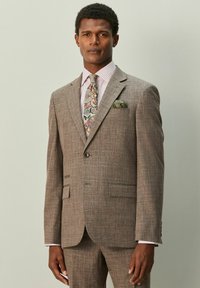 Brauner Karoanzug mit Spitzklappen, zwei Vordertaschen und einer passenden Krawatte mit buntem Paisley-Muster. Weißes Hemd mit rosa Streifen.