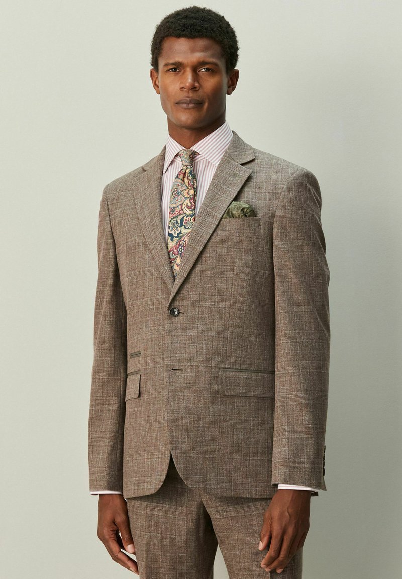 Brauner Karoanzug mit Spitzklappen, zwei Vordertaschen und einer passenden Krawatte mit buntem Paisley-Muster. Weißes Hemd mit rosa Streifen.