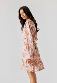 La robe présente un motif floral en paisley rose sur un fond clair. Manches longues et ceinture à nouer à la taille, avec un ourlet à volants. Tissu doux.