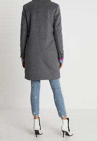 Šedý vlněný kabát s přímým střihem, zdobený knoflíky a barevnými manžetami, doplněný svetle modrými zkrácenými džínami s hvězdným vzorem a bílými podpatky.