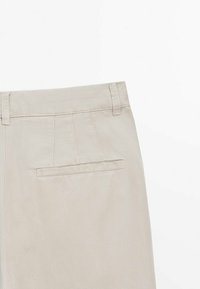 Pantalon beige à la texture lisse, doté d'un pli devant, d'une ceinture cousue et d'une poche arrière unique. Design simple et ajusté.