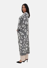 Cardigan lungo grigio a maglia con un motivo a stampa leopardata bianca, orlo a coste, maniche lunghe e tasche laterali. Indossato sopra pantaloni chiari.