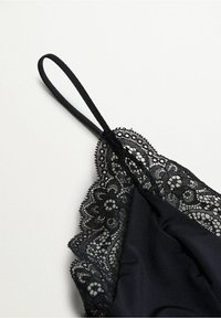 Top noir à fines bretelles spaghetti avec bordure en dentelle florale le long du décolleté et des bords, présenté sur un fond clair uni.