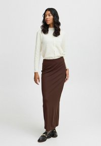 Blusa a maniche lunghe in texture crema abbinata a una gonna marrone aderente. Scarpe nere con accessori in oro. Tessuto liscio, silhouette elegante.