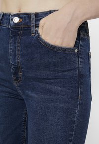Jeans de mezclilla azul oscuro con costuras naranjas en contraste, diseño de cintura alta y un bolsillo frontal. Cierre con botón y cremallera visible.