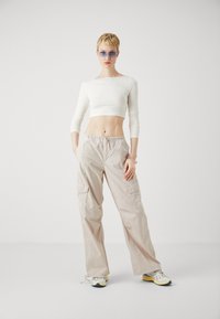 Top alb cu mâneci lungi, tăiat, pantaloni cargo bej cu buzunare și pantofi sport deschisi. Modelul poartă ochelari de soare cu nuanță albastră și o brățară.