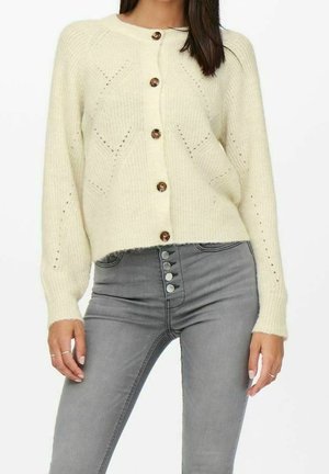 Cream knitted cardigan with a round neck, button closure, and openwork details. Paired with high-waisted grey jeans featuring snap buttons.