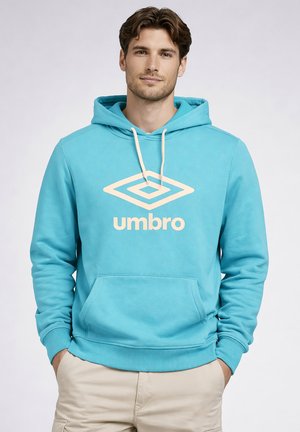 Homme portant un sweat à capuche bleu clair Umbro avec poche avant et pantalon beige, mains dans les poches, face à l'avant sur un fond uni.
