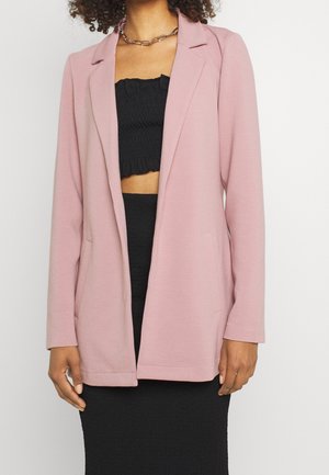 Vrouw draagt een lichtroze blazer over een zwarte buisvormige top en zwarte rok, met een kettingketting, tegen een effen witte achtergrond.