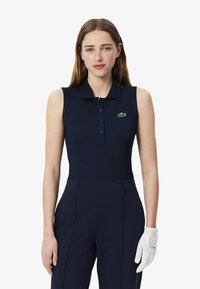 Polo sans manches bleu marine avec un col et une patte de boutonnage, arborant un petit logo de crocodile vert. Porte avec un pantalon bleu marine et un gant de golf blanc.